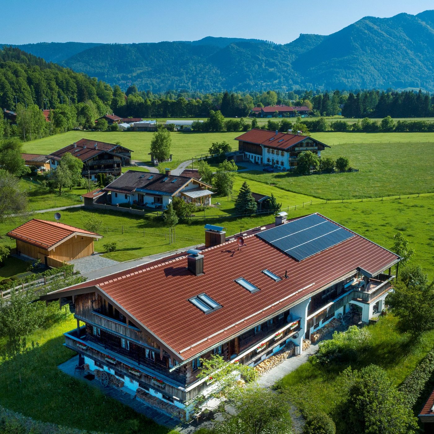 Luftbild: der Gasperhof mit Solardach, dahinter Wiesen, Höfe und die Berge des Tegernseer Tals